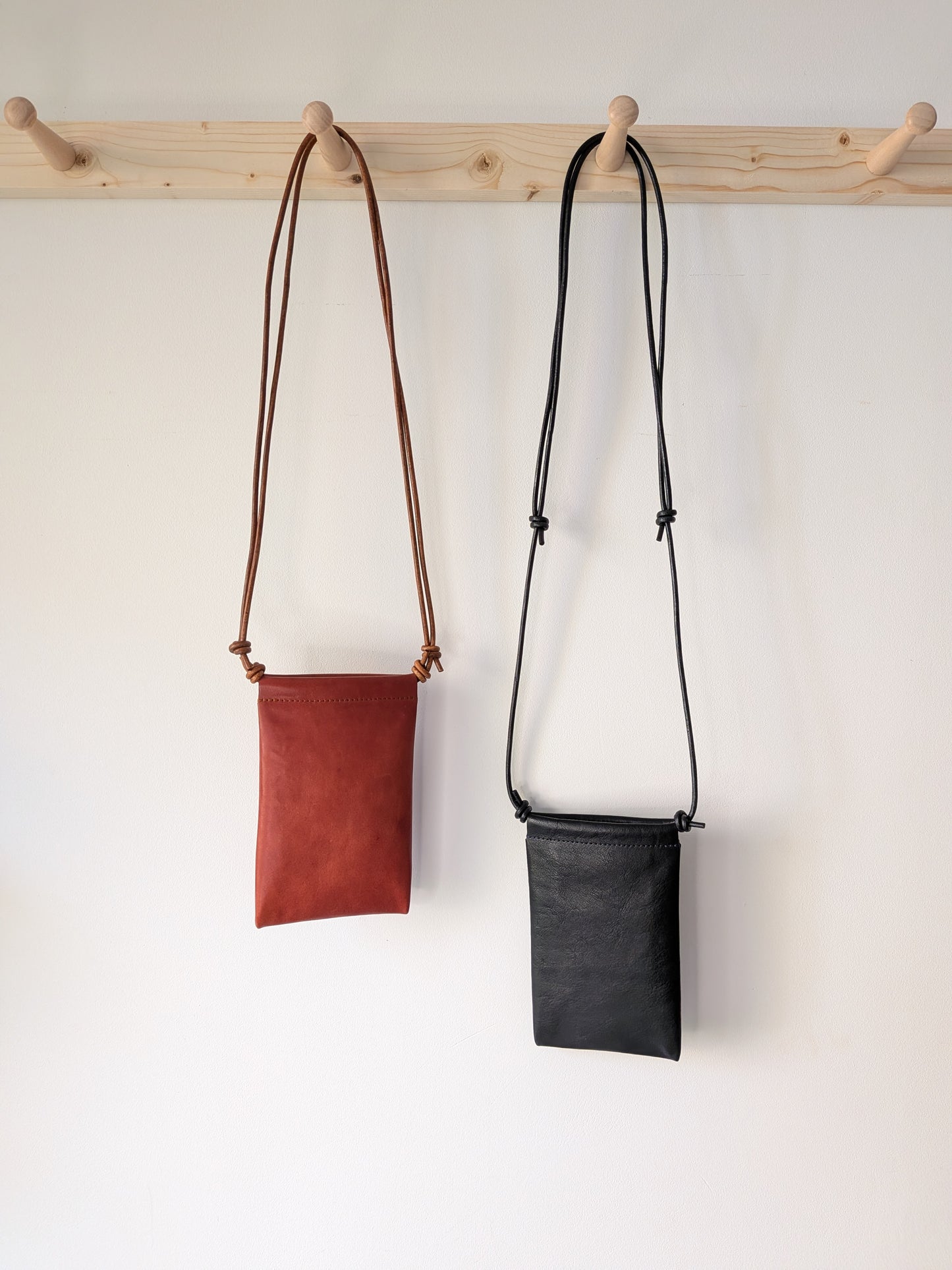 Two leather crossbody phone bags, one brow and one black, hanging on a wooden rack against a white wall.