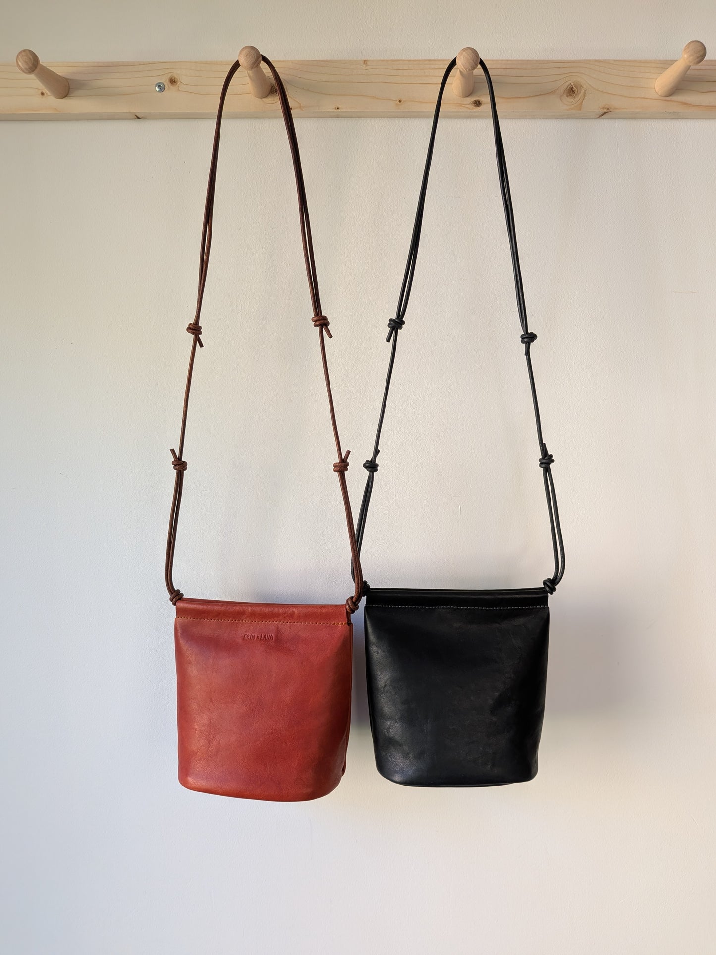 Two leather bags, one red and one black, hanging on a wooden rack against a white background.
