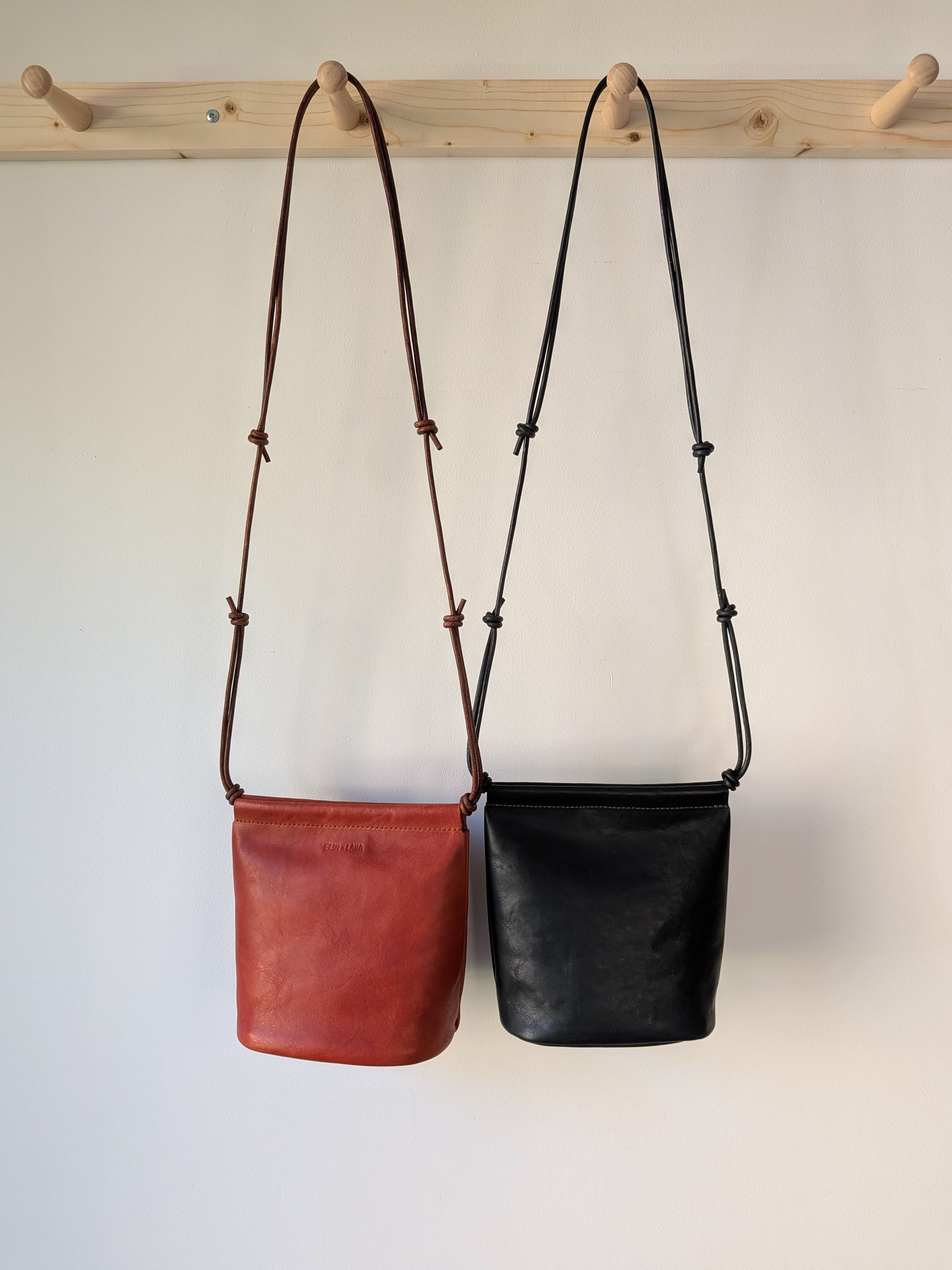 Two leather bags, one red and one black, hanging on a wooden rack against a white background.