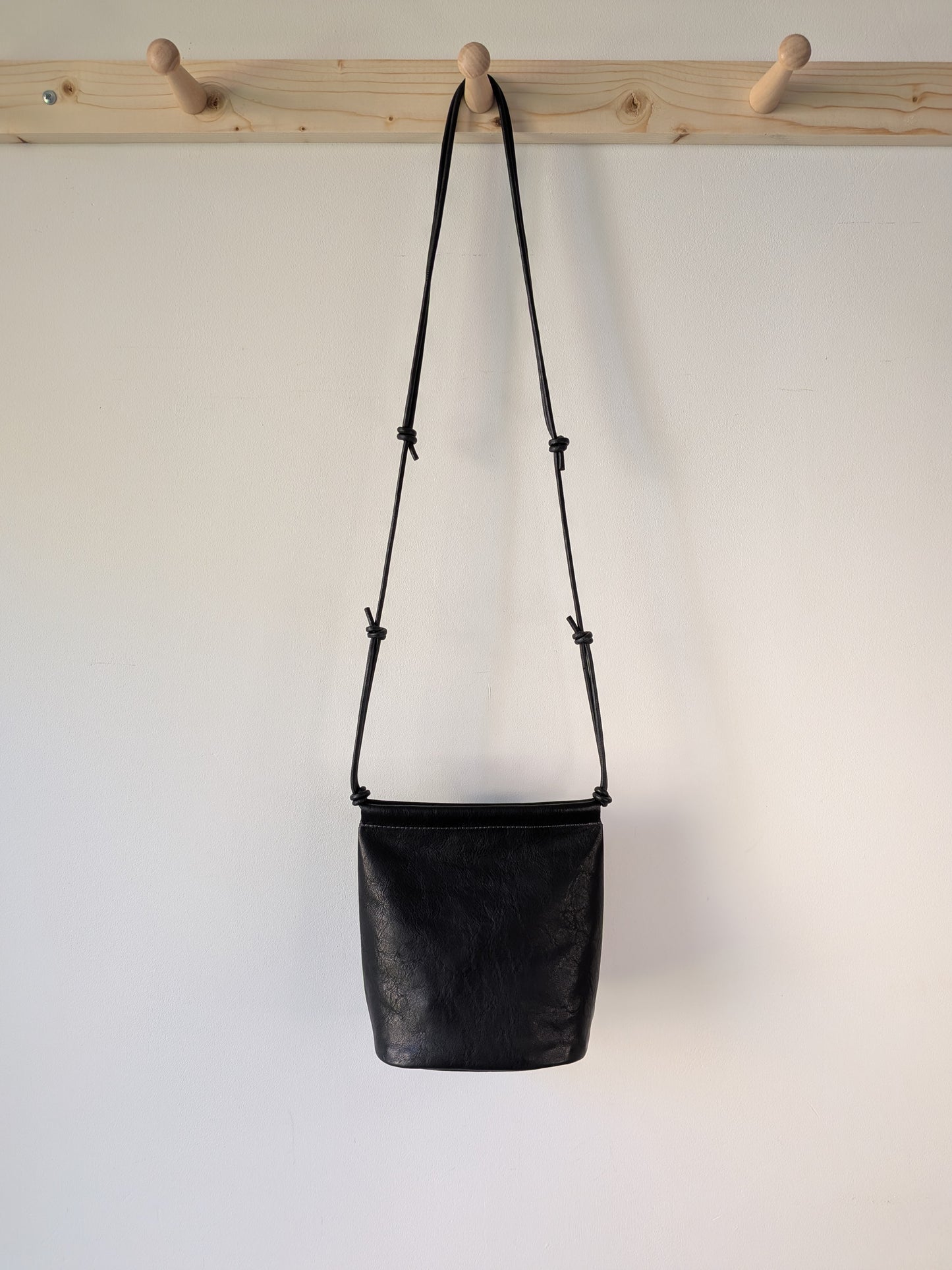 Black leather bag with a strap hanging on a wooden rack against a white background