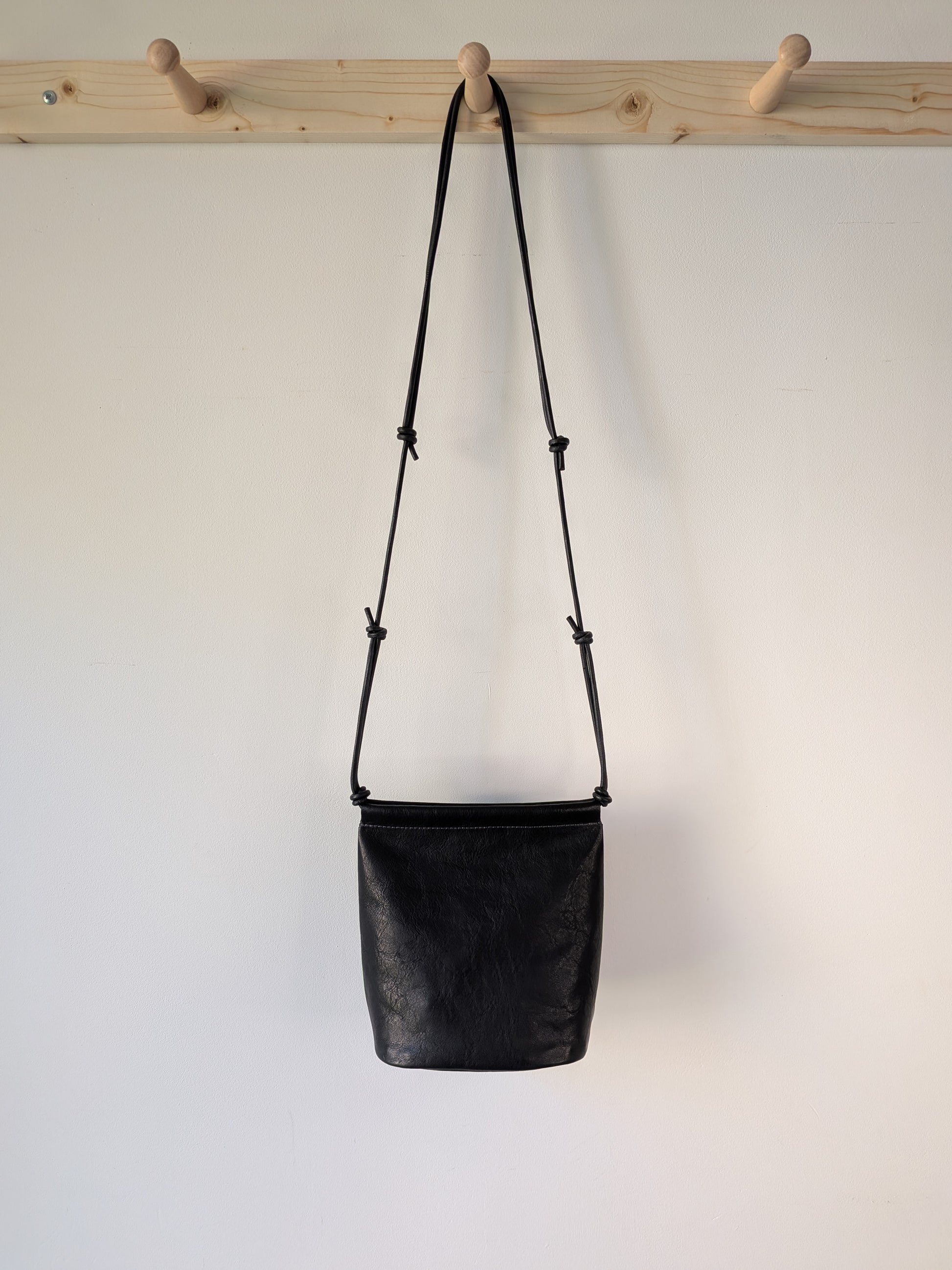 Black leather bag with a strap hanging on a wooden rack against a white background