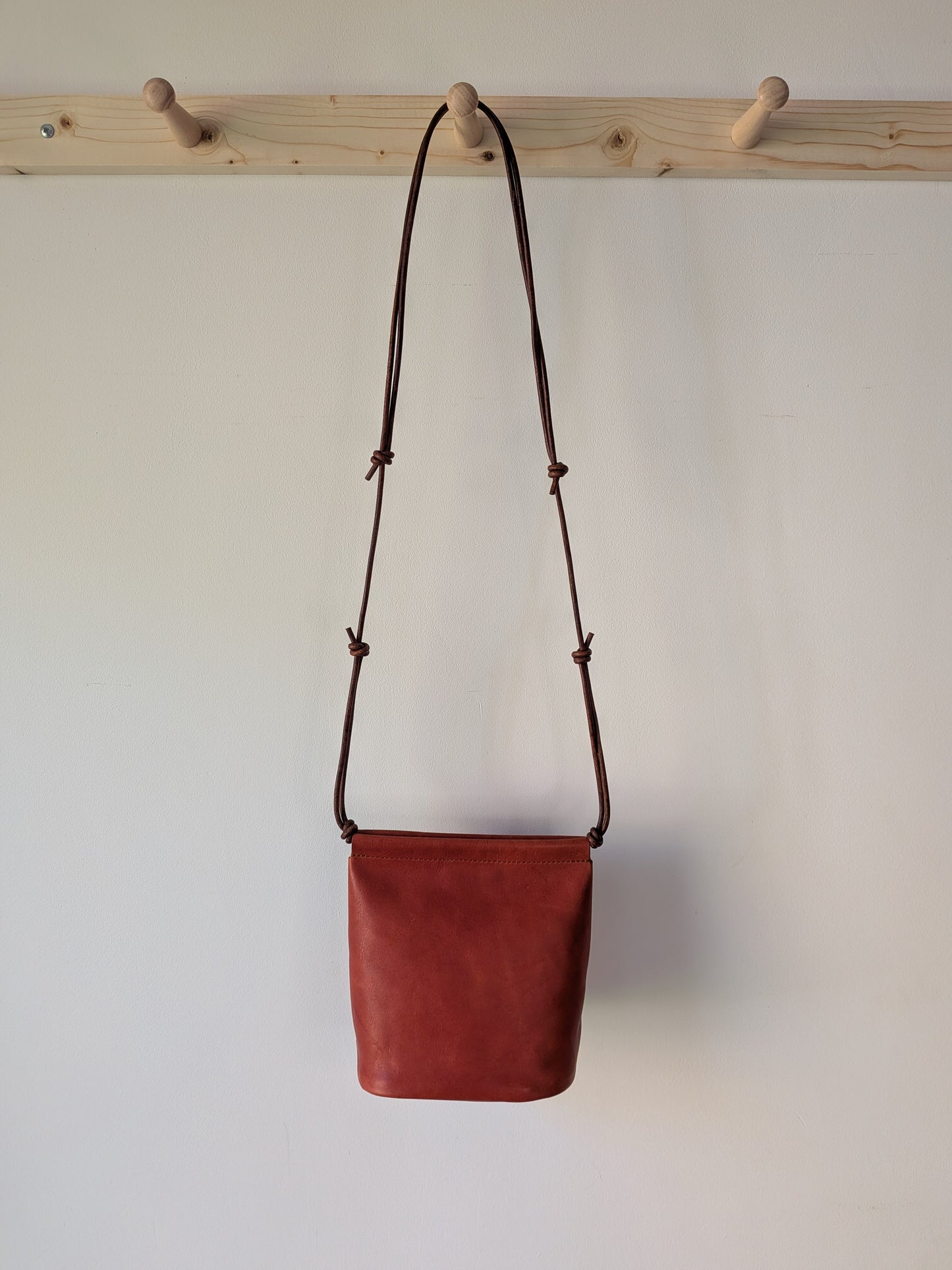 Red leather bag with a brown strap hanging on a wooden rack against a white wall.