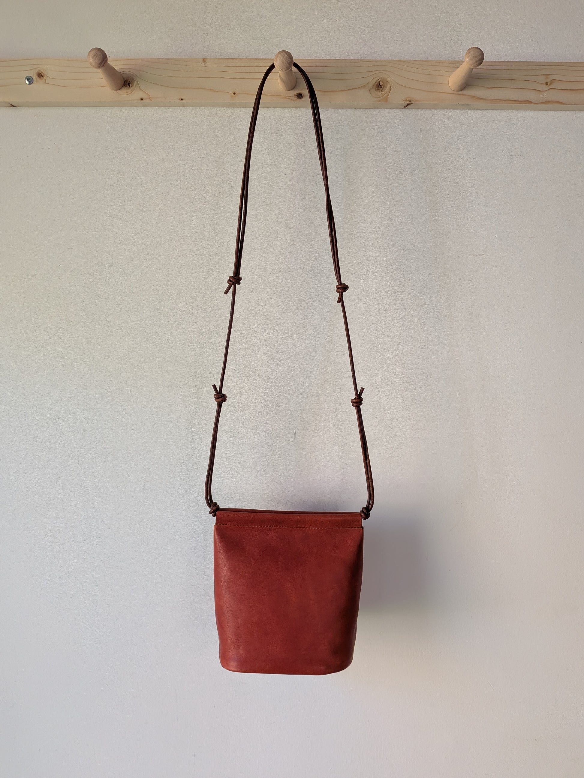 Red leather bag with a brown strap hanging on a wooden rack against a white wall.